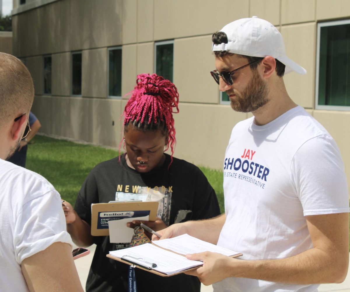 Shooster, who is wearing a white shirt representing his campaign for state representative, holds a clipboard while a student fills out a voter registration form.